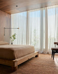 A minimalist bedroom with a wooden bed frame, light brown bedding, and a small plant on the nightstand. Floor-to-ceiling sheer curtains filter soft daylight. A simple chair sits near the window on the Armadillo&Co Meadow Rug - Brick.