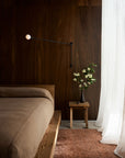 A minimalist bedroom features wood-paneled walls, a bed with brown bedding, a wooden side table topped with white flowers, and the Armadillo&Co Meadow Rug - Brick bringing retro 70s charm under sheer white curtains that filter natural light.