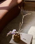 A small ceramic vase with delicate purple flowers and a long branch sits on a light table beside a curved brown sofa and the Petra Rug - Blanc by Armadillo&Co in a sunlit room.