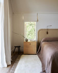 Minimalist bedroom with a beige bed, brown blanket, light wood nightstand, small vase with branches, dark stool, Armadillo&Co Petra Rug in Blanc, and sunlight streaming through a window with a white shade and greenery outside.