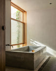 Minimalist bathroom with a large window, concrete soaking tub, and candle on the ledge. Natural light fills the space, greenery by the window, and an Armadillo&Co Willow Rug - Cypress woven from jute and wool lies on the floor.