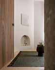 Minimalist interior featuring a textured beige wall, wooden door, Armadillo&Co Willow Rug - Cypress, small round table with teapot and cup, recessed niche with plant, and abstract white artwork on the wall.