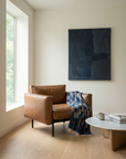 A modern brown leather armchair sits by a large window, draped with the Indigo Horizon — Vintage Baule Cloth Throw by Riad Jardin Des Biehn. Nearby, a round coffee table holds a mug and book beneath a dark abstract painting.