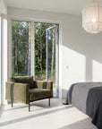 A modern bedroom with a large windowed door overlooking a green forest. Sunlight fills the space, which features Wooden Stories' Attendant Lounge Chair | Fabric, a gray bedspread, light wood floors, and a white pendant lamp.