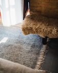 A cozy corner with a furry beige bench, the HIMALAYAN HERMITAGE Radhi Rug - Silver (hand-woven wool with fringed edges), sunlight streaming through a window, and a rustic brick wall in the background.