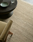 A close-up of the textured beige Mojave Rug - Cashew by Armadillo&Co, paired with a brown round table, green plate, glass of water, and a tan upholstered chair in a minimalist modern living space.