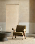 A modern olive-green armchair with wooden legs sits on an Armadillo&Co Mojave Rug in Cashew, beside a round dark wood coffee table with a glass carafe, set against a minimalist beige wall.