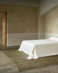 Minimalist bedroom with a simple bed covered in a white sheet, set on the green Mojave Rug – Laurel by Armadillo&Co. The room features beige walls, concrete floors, and an open wooden door on the left.
