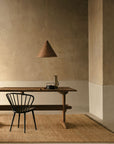A minimalist dining area with a wooden table, black chair, woven pendant, and simple tableware sits on the Armadillo&Co Mojave Rug – Natural in a hand-knotted Tibetan weave, set against a textured beige wall.