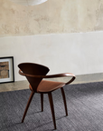 A wooden chair with curved armrests sits on the Sherpa Rug in Charcoal by Armadillo&Co in a minimalist room. A framed picture leans against the wall, and a paper lantern hangs from the ceiling above.