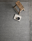 A small wooden stool and a beige book topped with a dark bowl sit on the Armadillo&Co Sherpa Rug in Pumice, with part of a speckled concrete floor visible at the edge.