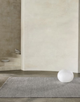 Minimalist room with cream walls, an Armadillo&Co Sherpa Rug in Pumice, a white spherical object, a small marble side table with a cup, and some decorative bowls on stone steps in the background.
