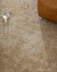 A glass of water sits on a light beige Armadillo&Co Sistine Rug - Manuka, beside the corner of a tan leather ottoman.