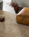 A tan carpeted floor with a large brown leather ottoman, a brown potted plant with dried branches, wooden blocks, and the Armadillo&Co Sistine Rug in Manuka creates a modern, minimalist room set against a striking concrete wall and floor.