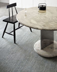 A round marble table with a metal pedestal base sits on the Corsica Rug - Capri by Armadillo&Co. A black wooden chair, glass cup, and black teapot are arranged beside and on the table in this modern, minimalistic room.