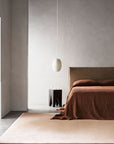 Minimalist bedroom with a brown bedspread, neutral walls, and the Armadillo&Co Eden Rug in Abalone. The space is finished with a dark side table and glassware, black bench, white pendant light, and natural light from a left-side doorway.