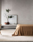 Minimalist bedroom in neutral tones featuring a wooden chair, a bed with tan bedding, the Eden Rug - Nougat by Armadillo&Co in hand-knotted wool, a potted plant on a low shelf, framed picture, and soft natural light from the doorway.
