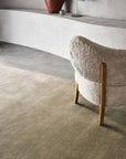 A curved, modern chair with soft white upholstery and wooden legs sits on the Armadillo&Co Eden Rug - Verona in a minimalist room with light walls and a built-in ledge holding a lamp and vase.