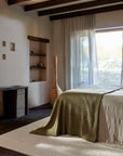 A serene bedroom with a neatly made bed in green and beige linens, a woven lamp, open shelves with decor, an Armadillo&Co Crest Rug - Poplin in a herringbone pattern, and sheer curtains letting sunlight in through a large window.