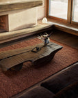 A rustic wooden coffee table sits on the Mason Rug - Sangria by Armadillo&Co, handwoven from New Zealand wool, in a sunlit living room with a low hearth and large window. On the table are a modern vase with flowers and a small teapot.