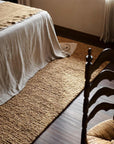 A cozy bedroom corner features a textured Mason Rug - Yolk by Armadillo&Co, a bed with a draped linen blanket, and a dark wooden chair with a woven seat, all softly lit by natural light from the window.