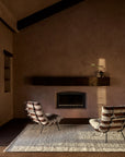 A minimalist living room features two striped lounge chairs on the Tempest Rug - Tundra by Armadillo&Co, made from New Zealand wool, facing a built-in fireplace with a wooden mantle holding a ceramic vase and greenery, lit by warm natural light.