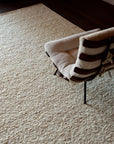 The Tempest Rug - Tundra by Armadillo&Co, a textured cream hand-knotted New Zealand wool rug, lies on a wooden floor beside a cushioned chair with wooden slats and cream upholstery.