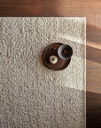The Tempest Rug - Tundra by Armadillo&Co, crafted from New Zealand wool in a textured beige finish, lies on a wooden floor with sunlight casting soft shadows; atop it sits a dark tray with a ceramic vase and bowl of nuts.