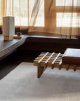 A modern, minimalist room features a wooden slatted bench with brown leather cushion, built-in concrete seating, books, sheer curtains, and the Armadillo&Co Thatch Rug - Dew in light beige 100% wool beneath large sunlit windows.