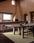 A rustic dining room with a large wooden table, simple chairs, an Armadillo&Co Thatch Rug - Marsh of wool fiber, stucco walls, arched windows, a built-in corner fireplace, all bathed in natural light.