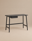 The Wooden Stories Author Desk in black oak features angled legs and a single central drawer, its sleek Danish-inspired lines highlighted against a plain beige background.