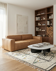 A modern living room features the Wooden Stories Baker Sofa in tan leather, a geometric rug, a white oval coffee table, and a built-in wooden bookshelf with books and firewood. Sunlight filters through sheer curtains.