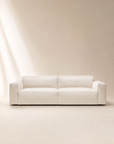 A modern Baker Sofa | Fabric by Wooden Stories, upholstered in cream fabric with wide armrests and a minimalist Danish design, features plush feather filling and is set against a neutral beige backdrop illuminated softly from the left.