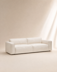 A modern, minimalist Baker Sofa | Fabric by Wooden Stories, featuring wide armrests and low backrests in a cream color, sits on a light beige background with soft natural lighting.
