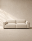The Baker Sofa | Fabric by Wooden Stories, featuring wide armrests and plush feather-filled cushions, sits against a beige wall and floor, illuminated softly from the left by natural light.