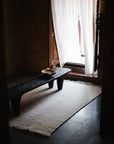 A minimalist room features a dark wooden bench on the Radhi Rug - Snow by HIMALAYAN HERMITAGE, with a wooden bowl atop the bench and sunlight streaming through sheer white curtains on an open window.