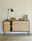 The Blackbird Sideboard by Ethnicraft, crafted from solid European oak and accented with sleek black legs, is styled with a black desk lamp, office supplies, and two folders arranged on its surface. One of its cabinet doors is slightly ajar, offering a glimpse of the items stored inside. This modern piece sits elegantly on a light wooden floor.