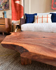 A close-up of the Bolinas Coffee Table - Claro Walnut by SOBU, handmade in Oakland, is shown in a cozy living room with a modern sofa, blue and white pillows, a wooden side table, and colorful wall art in the background.