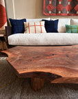 A close-up of the SOBU Bolinas Coffee Table in Claro Walnut, handmade in Oakland, is shown in front of a white sofa with plaid throw pillows. A patterned rug and wall hanging complement the modern setting.
