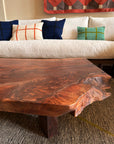 A close-up of the Bolinas Coffee Table - Claro Walnut by SOBU, handmade in Oakland with a natural live edge, is set before a white sofa with colorful plaid pillows, cozy textured fabrics, and a patterned wall hanging.