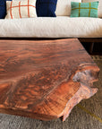 A close-up of the Bolinas Coffee Table - Claro Walnut by SOBU, featuring a rustic live edge. A beige sofa with colorful plaid cushions is behind it. Handmade in Oakland, it adds modern style to any space.