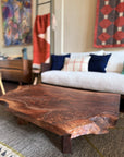 The Bolinas Coffee Table - Claro Walnut by SOBU, featuring natural edges, sits on a patterned rug in a cozy living room with a white sofa, pillows, colorful blankets, and vibrant artwork—expertly handmade in Oakland.