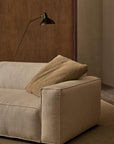 The Baker Modular Sofa | Fabric • M9 by Wooden Stories, in modern beige, features a large brown cushion and sits on a textured rug beneath a black minimalist wall lamp, set against a brown wooden panel and taupe wall.