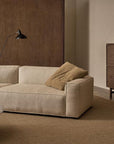 A minimalist living room with the Baker Modular Sofa | Fabric • M9 by Wooden Stories, a tan throw blanket, a round marble coffee table, wooden cabinet, brown paneled walls, and a wall-mounted black lamp.