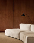 A modern, minimalist living room showcases the customizable Baker Modular Sofa | Fabric • M9 by Wooden Stories on a textured rug, set against a smooth brown wooden wall with a black adjustable wall-mounted lamp above.