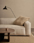 A minimalist living room features the Baker Modular Sofa | Fabric • M9 by Wooden Stories in beige, a textured tan cushion, wooden coffee table with a small cup, woven rug, and a black wall lamp set against a neutral brick wall.