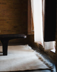 A wooden bench sits on the Radhi Rug - Snow by HIMALAYAN HERMITAGE, a hand-woven wool rug inspired by Nepali design, beside a window with sheer white curtains that allow natural light and soft shadows into the room.