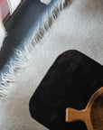 A close-up of the HIMALAYAN HERMITAGE Radhi Rug - Snow, a light-colored hand-woven wool rug with fringe, sits by a window with sheer white curtains. A wooden tray and carved bowl rest on the rug.