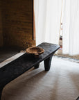 A dark wooden bench with a carved bowl on top sits on the Radhi Rug - Snow by HIMALAYAN HERMITAGE in a room with brick walls and white curtains letting in soft natural light.