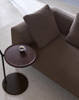 A brown sofa with two cushions sits beside the Wooden Stories Bystander Side Table, which holds a small cup of espresso. The scene features a light rug and wooden flooring in a minimalist room.